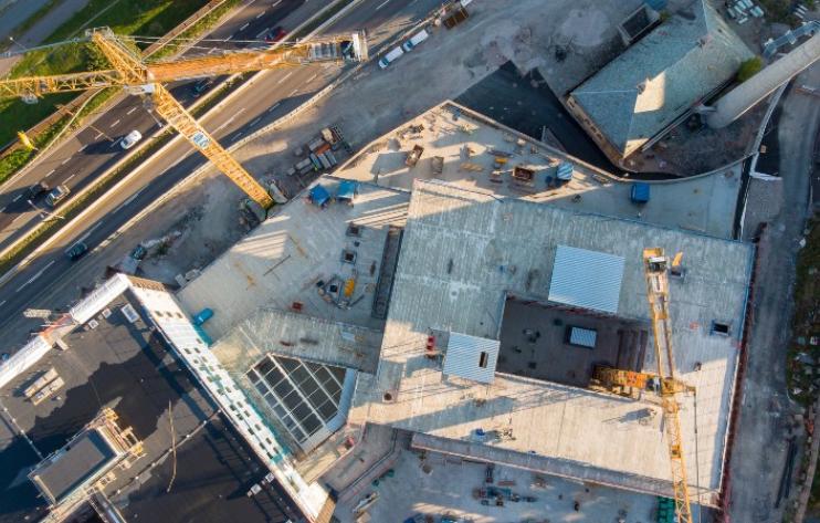 En flygbild som visar en byggarbetsplats ovanifrån med pågående arbete, byggkranar och en intilliggande väg med trafik.